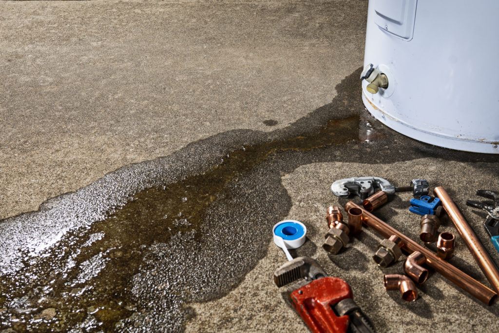 Water leaking from the plastic faucet on a residential electric water heater sitting on a concrete floor with a red pipe wrench, tubing cutters, teflon tape and copper fittings in the foreground to repair or place the appliance.